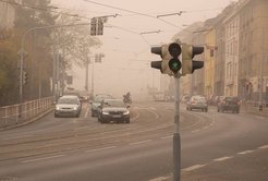 A street with a green traffic light in foggy conditions, featuring several cars, tram tracks, and buildings lining both sides of the road.