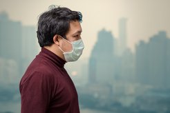 A man in a dark red sweater and face mask stands in front of a hazy cityscape, with tall buildings partially visible in the background.