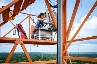 High above the jungle: Atmospheric chemist Nora Zannoni checks her instruments at the Amazon Tall Tower Observatory ATTO. Source: Fabio Cian Researcher Nora Zannoni sits on a high scaffold on the ATTO research tower, operating technical equipment high above the Brazilian rainforest.