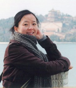 Researcher Yafang Cheng, wearing a brown jacket and gray scarf, stands in front of a lake with a temple in the background.