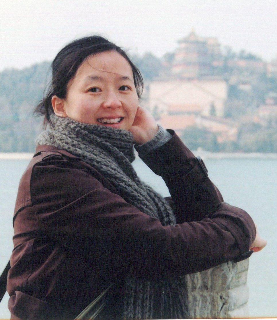 Researcher Yafang Cheng, wearing a brown jacket and gray scarf, stands in front of a lake with a temple in the background.